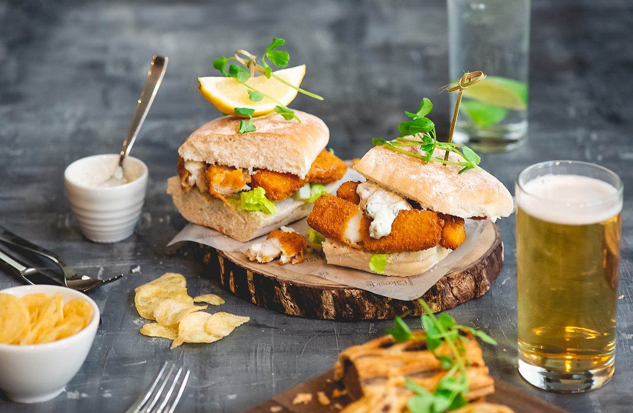 Fish sandwiches stack with fried fillets, lettuce, and tartar sauce, topped with lemon and greens; presented on a rustic board beside potato chips, a dip cup, cutlery, and a glass of beer.